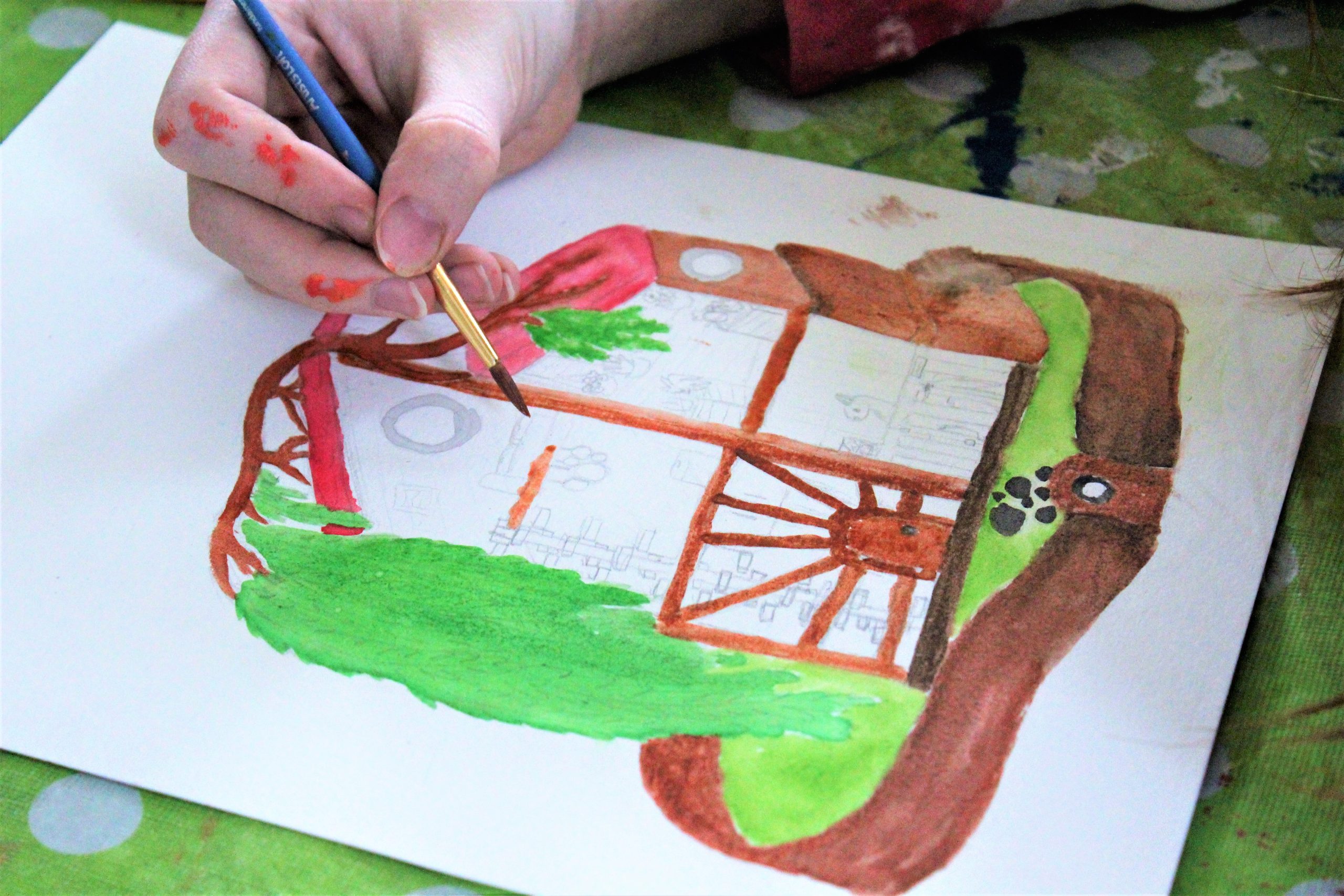 A photograph of an individual painting a brown house with green leaves while being on top of a green and white sheet.