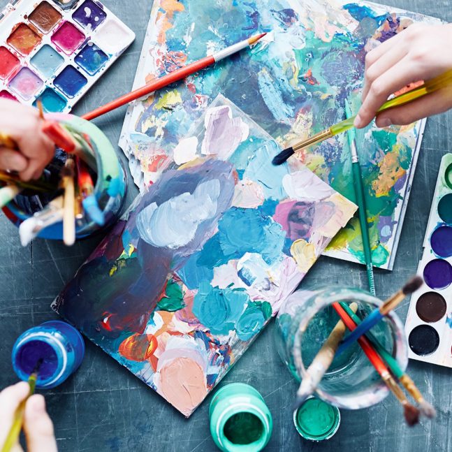 people's hands painting surrounded by paint palettes, brushes, and painted paper