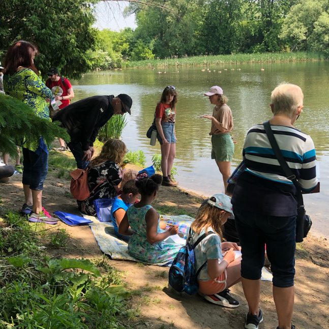 a group of adults and children sitting and standing at the bank of a river, surrounded by greenery on a sunny day