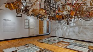 an art installation in a gallery consisting of a variety of wooden chairs suspended from the ceiling, with many mirrors arranged on the floor below. Wooden doors are installed on the wall in the background next to text