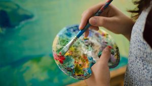a person's hands dipping a paintbrush into red paint on a palette with lots of paint on it
