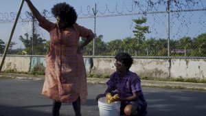two women on a road with barbed wire fence and vegetation in the background. The woman on the left stands and holds a long pole in her hand while the other squats by a bucket and holds fruits in her hands