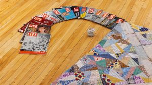 an art installation with a semi-circle of "Life" magazines on the floor next to a quilt and a stone with a ceramic lid