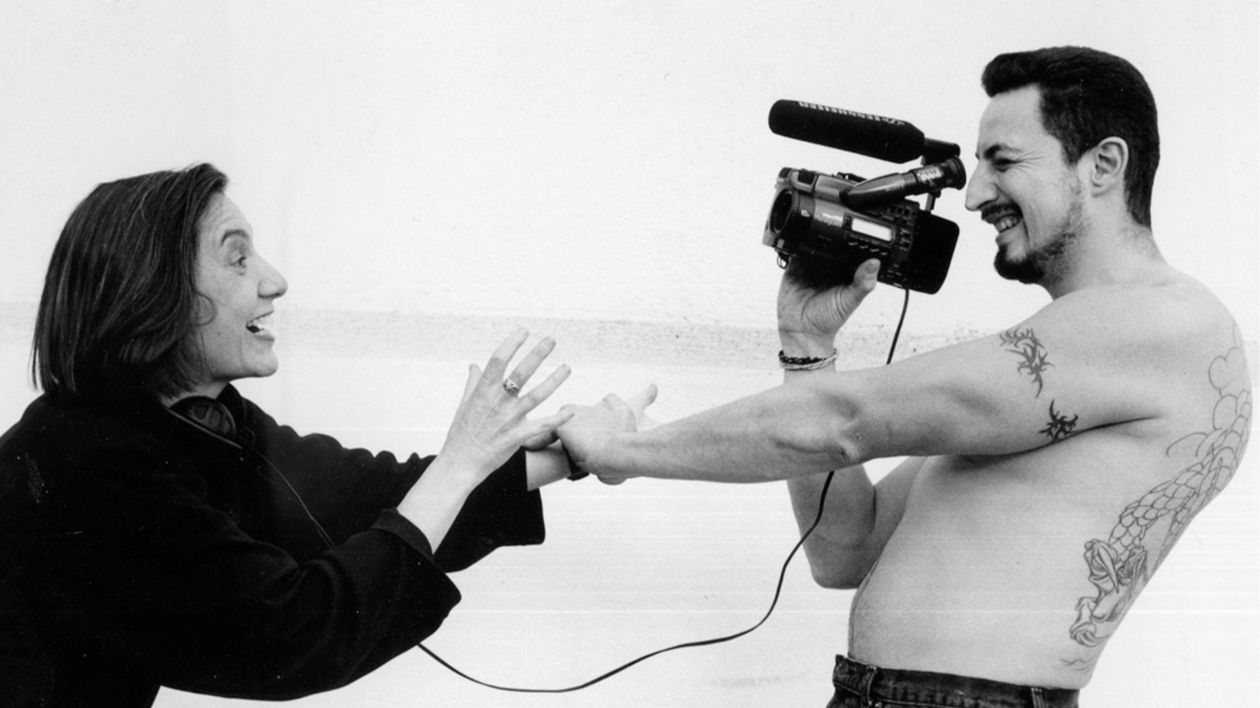 A candid black-and-white photograph of two people smiling and holding hands. The person on the right, who appears to be filming the other person with a camcorder, has the camcorder raised to their eye.